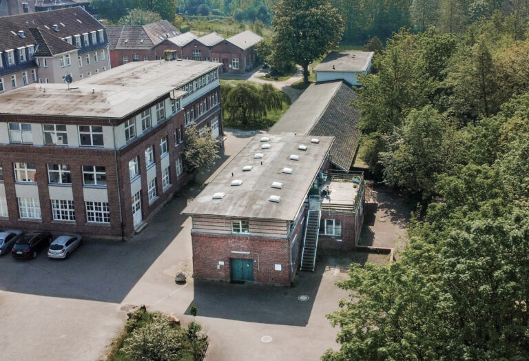 Luftaufnahme eines kleinen Backsteingebäudes mit einer Treppe an der Seite, das an ein größeres Gebäude in einem von Grün umgebenen Komplex von Hartig & Hirsch angrenzt. In der Nähe der Gebäude sind mehrere geparkte Autos zu sehen, die eine durchdachte Standortentwicklung veranschaulichen.