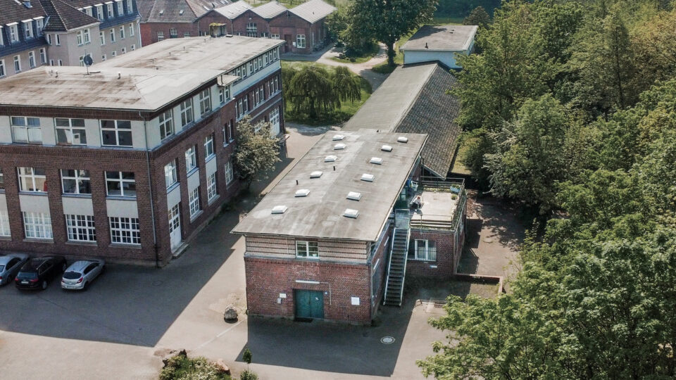 Luftaufnahme eines kleinen Backsteingebäudes mit einer Treppe an der Seite, das an ein größeres Gebäude in einem von Grün umgebenen Komplex von Hartig & Hirsch angrenzt. In der Nähe der Gebäude sind mehrere geparkte Autos zu sehen, die eine durchdachte Standortentwicklung veranschaulichen.