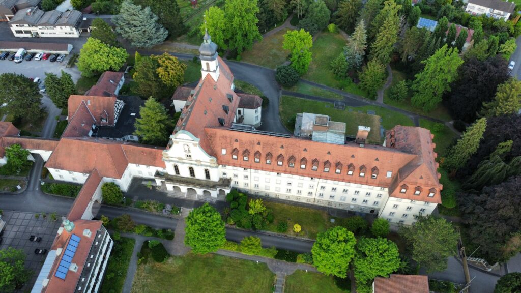 Luftaufnahme des Klosters Maria Hilf, einem großen, historischen Gebäude in Bühl mit Ziegeldach, zahlreichen Fenstern und einem Uhrturm. Umgeben von Grün befindet sich dieses transformative Gebäude in einer gepflegten Umgebung mit anderen Gebäuden in der Nähe.