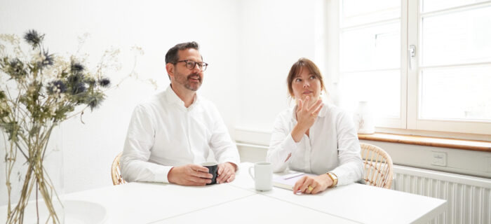 In einem hellen Raum mit Fenster und einer Vase mit Blumen sitzen zwei Menschen an einem weißen Tisch. Beide tragen weiße Hemden und haben jeweils eine Tasse vor sich. Sie diskutieren über Standortentwicklungsstrategien für Hartig & Hirsch.