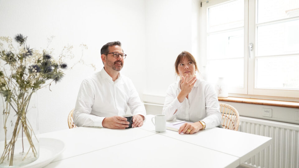In einem hellen Raum mit Fenster und einer Vase mit Blumen sitzen zwei Menschen an einem weißen Tisch. Beide tragen weiße Hemden und haben jeweils eine Tasse vor sich. Sie diskutieren über Standortentwicklungsstrategien für Hartig & Hirsch.