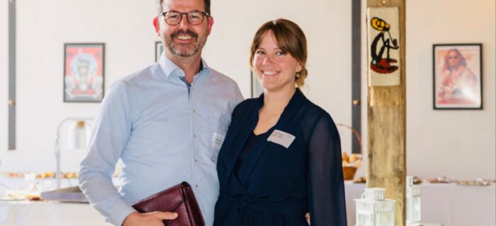 In einem gut beleuchteten Raum in Dormagen stehen ein Mann und eine Frau lächelnd. Der Mann hält ein kastanienbraunes Notizbuch in der Hand und beide tragen Namensschilder. Kunstwerke erhellen die Wände hinter ihnen.