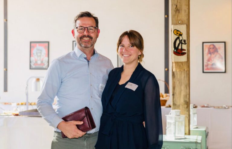 In einem gut beleuchteten Raum in Dormagen stehen ein Mann und eine Frau lächelnd. Der Mann hält ein kastanienbraunes Notizbuch in der Hand und beide tragen Namensschilder. Kunstwerke erhellen die Wände hinter ihnen.
