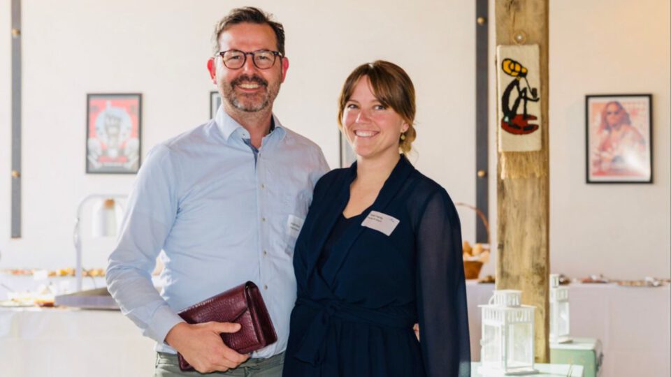 In einem gut beleuchteten Raum in Dormagen stehen ein Mann und eine Frau lächelnd. Der Mann hält ein kastanienbraunes Notizbuch in der Hand und beide tragen Namensschilder. Kunstwerke erhellen die Wände hinter ihnen.